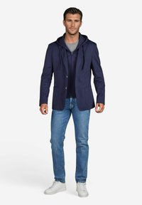 Navy jack met een capuchon, twee voorzakken, gecombineerd met een grijs shirt en lichtblauwe jeans. Witte sneakers maken de outfit compleet.