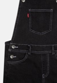 Černé denimové lacláče s bílým šitím, kovovými prvky, přední kapsou a nastavitelnými popruhy. Klasický design pro dlouhou životnost.