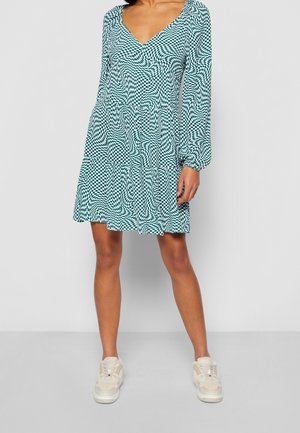 Frau trägt ein langärmliges, grün-weiß kariertes Kleid mit tiefem V-Ausschnitt und weißen Turnschuhen, steht auf einem grauen Boden.