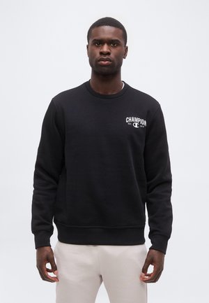 Mand iført sort Champion-sweatshirt med hvidt logo på brystet, stående mod en ensartet lysegrå baggrund.