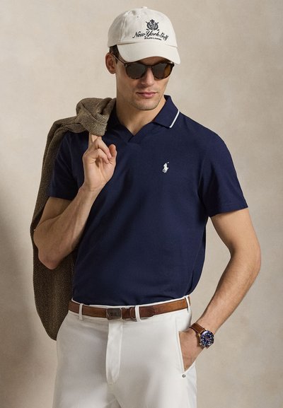 Άνδρας με μπλε πόλο, λευκό καπέλο με την επιγραφή "New York Golf", γυαλιά ηλίου, λευκό παντελόνι, καφέ ζώνη, ρολόι και κρατώντας καφέ σακάκι στον ώμο.