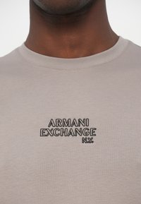 Light beige cotton t-shirt features a crew neck and embroidered "ARMANI EXCHANGE N.Y." logo in black. Smooth texture with minimal detailing.
