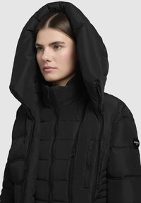 Svart puffjacka med en stor huva, dragkedja fram, quiltad textur och logotypapplikation på överarmen.