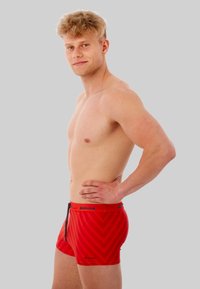 Shorts de bain rouges avec un motif en chevron, en tissu lisse. Présentent une ceinture avec cordon de serrage et un petit logo sur le devant.