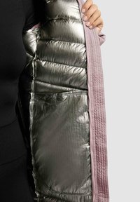 Hånd der holder en åben lys pink frakke, der afslører et sølvmetallic polstret interiør med kviltede lommer og sydet detaljer.