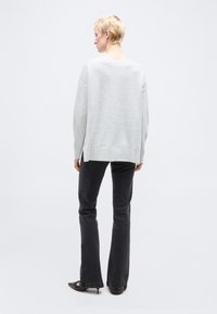 Άτομο με κοντά ξανθά μαλλιά, που φοράει άνετο ανοιχτό γκρι πουλόβερ, μαύρο καμπάνα παντελόνι και μαύρες μυτερές γόβες, που στέκεται με την πλάτη.