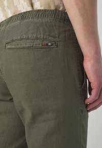 Maslinasto zelene hlače od lagane tkanine, s stražnjim džepom s višebojnim detaljem. Elastična naramenica za udobnost.
