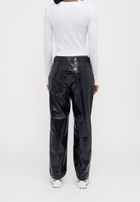 Person klädd i glänsande svarta faux läderbyxor med krokodilmönster, vit långärmad crop top och vita sneakers, vänd bort.