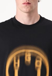Sort bomulds T-shirt med et stort gult grafisk print i et prikket mønster, centreret på brystet, med en rund halsudskæring.