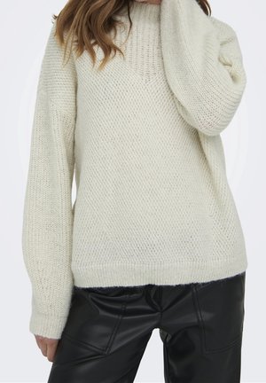Frau trägt einen cremefarbenen grobgestrickten Pullover mit hohem Kragen und schwarze Lederhose, posiert mit einer Hand nahe am Gesicht.