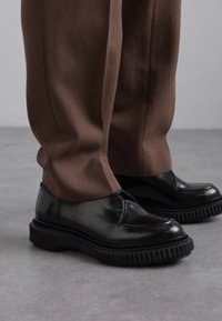 Schwarz polierte Lederschuhe mit einem eleganten Design, dicker gerippter Gummisohle und ohne sichtbare Schnürsenkel, kombiniert mit braunen Hosen.
