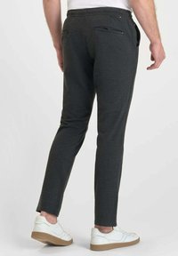 Man in een donkergrijze slim-fit broek met een achterzak met rits en witte sneakers, staand tegen een witte achtergrond.