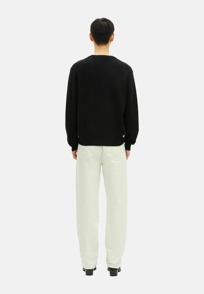 Mężczyzna stojący plecami, ubrany w czarną bluzkę z długim rękawem, jasne spodnie z tylnymi kieszeniami i czarne buty na jednolitym tle.