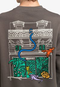 Sudadera gris que presenta un gráfico detallado de un edificio con un balcón, decorado con animales coloridos, plantas y una tabla de surf.
