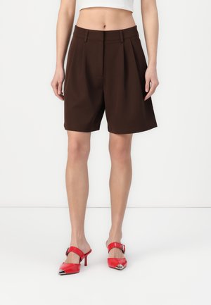 Shorts marron ajustés avec plis et taille mi-haute. Portés avec des chaussures à talons vernis rouges, présentant des embouts en argent et une accent de boucle.