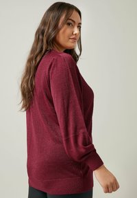 Pull en Bourgogne avec un design en tricot texturé, des manches longues, un ourlet côtelé et un subtil éclat. Porté sur des leggings noirs, présenté de côté.
