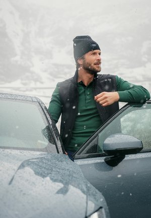 Mann in einer schwarzen Weste über einem langen grünen Shirt, trägt eine schwarze Mütze, steht neben einem regennassen dunkelgrauen Auto. Schnee fällt.