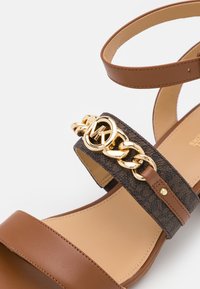 Sandale en cuir marron avec bride à la cheville et bande sombre à motifs ornée d'une chaîne dorée et du logo MK.