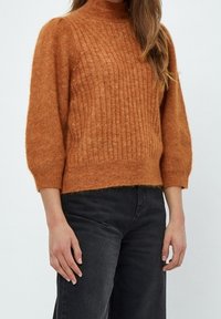 Mujer vistiendo un suéter de punto acanalado color óxido con mangas tres cuartos y unos vaqueros negros de tiro alto contra un fondo plano.