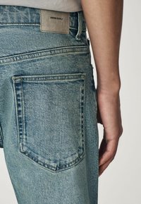Jean en denim bleu clair avec une finition texturée, deux poches arrière et une étiquette en cuir avec "DENIM SUPPLY" cousue sur la ceinture.