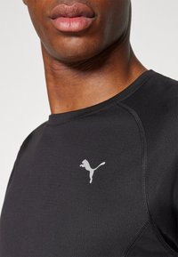 Schwarzes Sportshirt aus dehnbarem Material, mit rundem Ausschnitt und kleinem grauen Puma-Logo auf der Brust.