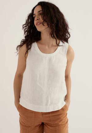 Femme aux cheveux bouclés et foncés, portant un haut blanc sans manches et un pantalon marron, debout les mains dans les poches, regardant légèrement sur le côté.