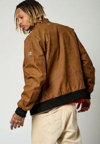 Veste bombardier en cuir marron avec une finition texturée, poignets noirs côtelés et un petit patch logo sur l'épaule. Portée par-dessus une chemise rouge.