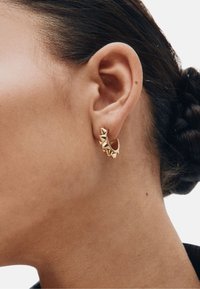 Kultainen rengaskoru, jossa on kaareva muotoilu ja pyramidinmuotoiset korostukset ulkoreunalla, teksturoitu pinta ja kiillotettu viimeistely.