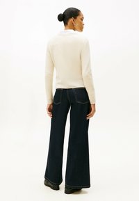 Vrouw met een bun, gekleed in een crème trui, witte blouse, donkere wijde jeans en zwarte schoenen, staande tegen een witte achtergrond.
