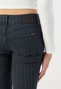 Persona vestida con jeans oscuros de rayas y una camiseta blanca de mangas largas con textura, mostrando el bolsillo trasero y la parte baja del torso.