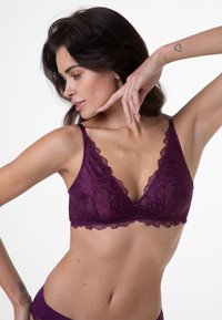Bralette in pizzo viola con scollatura a V profonda, bordi scallopati e spalline sottili. Il tessuto presenta un motivo floreale con una texture semi-trasparente.