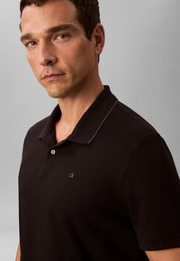 Mand med kort mørkt hår iført en sort polo med et lille "ck"-logo på brystet mod en ensartet grå baggrund.