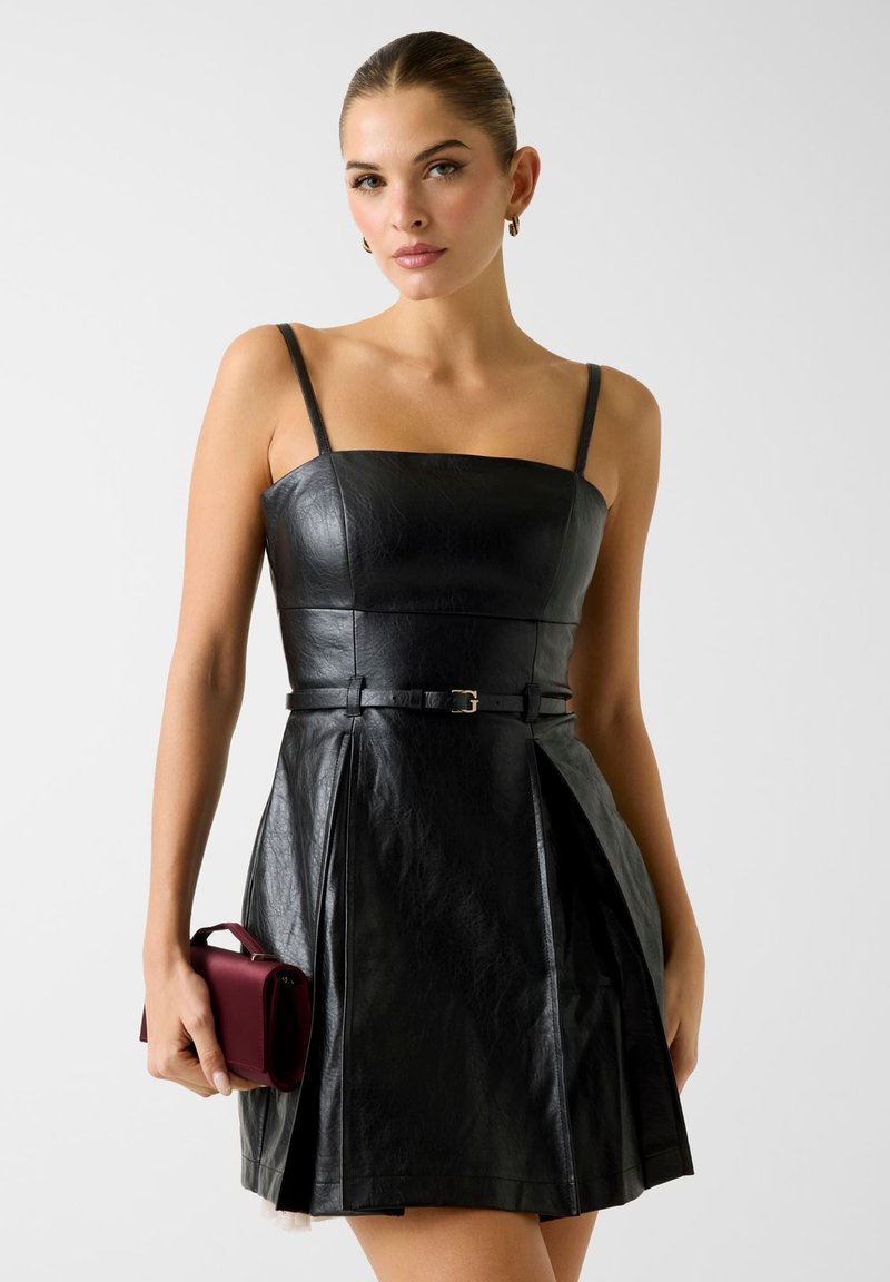 Robe en cuir noir avec un décolleté carré, des bretelles fines, une taille ceinturée et une jupe plissée. Le mannequin tient un petit sac à main bordeaux.