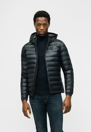 Giovane uomo che indossa una giacca imbottita nera con cappuccio, maglione nero a collo alto e jeans blu scuro, in piedi su uno sfondo chiaro e uniforme.