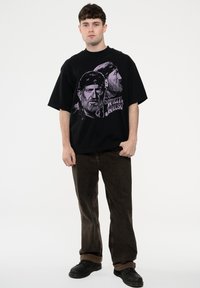 Black oversized t-shirt featuring a graphic print of two bearded men, with visible stars and design accents, paired with brown pants and black shoes.