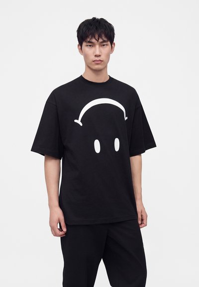 Jeune homme portant un T-shirt noir oversize avec un motif abstrait de visage blanc et un pantalon noir, debout devant un fond clair uni.
