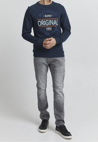 Marineblauwe crewneck sweater met meerkleurige opdruk met de tekst "ORIGINAL 1993," gecombineerd met lichtgrijze jeans en zwarte sneakers.