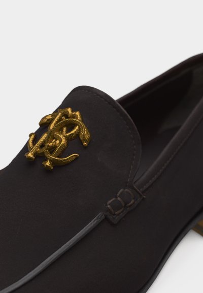 Μαύρο μοκασίνη από σουέντ με χρυσό έμβλημα λογότυπου που συνδέεται στενά στο πάνω μέρος και λεπτομερείς ραφές κατά μήκος των άκρων.