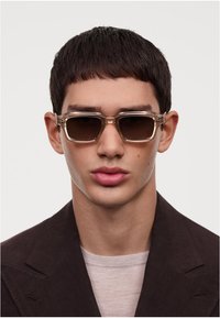 Ochelari de soare dreptunghiulari cu un cadru transparent și lentile închise la culoare. Cadru are accente argintii. Purtați de un model cu păr scurt și închis la culoare.