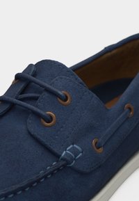 Κοντινό πλάνο ναυτικού μπλε μοκασίνι από σουέντ με δερμάτινα κορδόνια περασμένα από χάλκινα κουμπώματα και λεπτομέρειες ραψίματος σε ανοιχτό μπλε.