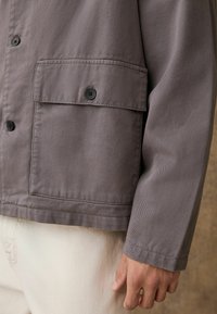 Szara bawełniana kurtka z zapięciem na guziki, z łatą kieszeni i teksturowanym materiałem. Rękaw ma starannie wykończony mankiet.