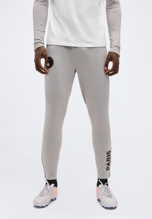 Mann trägt hellgraue Sporthose mit Paris-Logo am linken Bein und einem runden Emblem am rechten Oberschenkel, kombiniert mit weißen Stollenschuhen.