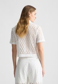Camicetta bianca corta con ricami a occhiello e ritagli circolari. Maniche corte, tessuto testurizzato e orlo pulito. Indossata con pantaloni abbinati.
