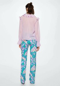 Jasnofioletowa przezroczysta bluzka z falbaniastym kołnierzykiem i mankietami, zestawiona z szerokimi spodniami w kolorowy, wirujący kwiatowy wzór w odcieniach zieleni, niebieskiego i kremowego.