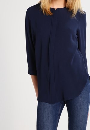 Woman wearing a navy blue long-sleeve blouse with a round neck and dark blue jeans, standing against a plain white background.