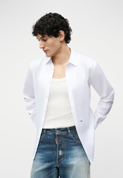 Jeune homme aux cheveux bouclés portant une chemise blanche ouverte sur un débardeur blanc et un jean bleu délavé, regardant vers le bas à gauche.