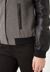 Chaqueta bomber de tela gris con mangas de cuero negro, puños acanalados y un bolsillo plano negro, con botones a presión y una superficie texturizada.