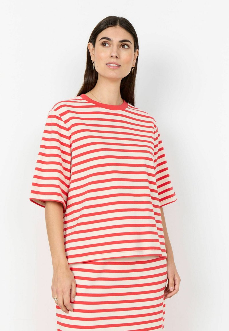 Femme aux longs cheveux foncés portant un haut à manches courtes rayé horizontalement rouge et blanc et une jupe assortie, debout devant un fond blanc.