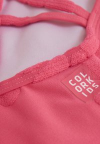 Rosa tyg med texturerade accenter och sömnad, som har en fastsatt etikett med texten "COLOR KIDS." Designen inkluderar en lagererad remdetalj.