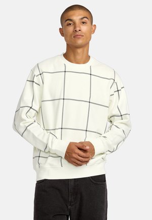 Sweatshirt crème clair avec un motif de grille noire. Doté d'un col ras du cou et de poignets côtelés, fabriqué en tissu doux. Porté avec un jean noir.